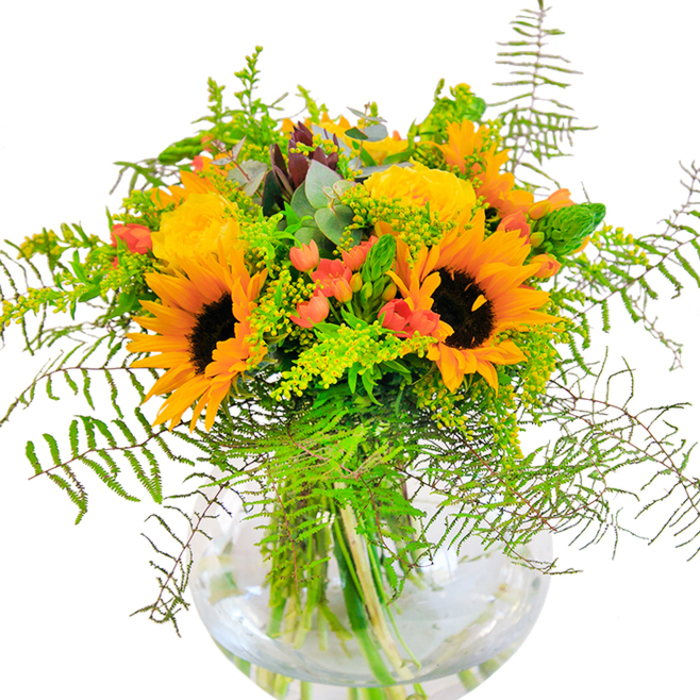 From the moment you glimpse this bouquet, a sense of sunlit abundance radiates forth, echoing the garden scenes found along East Finchley's tree-lined avenues. The clear, spherical glass vase cradles an arrangement where the joyful faces of sunflowers beam in golden glory, each dark brown center creating a dynamic focus amid a halo of orange-yellow petals. Sitting beside them, yellow garden roses spiral open, their plush, layered petals like the gentle folds of a newly blossoming rose in a local allotment. Among the blooms, clusters of red Hypericum berries shine, creating lively, jewel-like highlights against a backdrop of greens from varied foliage. Diameters of feathery ferns and sprigs of solidago-possibly tipped with tiny yellow flowers-extend outward, enhancing the bouquet's sense of organic movement, just as brambles and wildflowers do in Cherry Tree Wood. Eucalyptus leaves, with their subtle blue-green shades and rounded shape, bring a modern, aromatic note to the composition. The vase's clarity reveals stems in water, reinforcing a feeling of freshness and care so valued by those in East Finchley. The arrangement is alive with gentle fragrance, uplifting softness, and a visual celebration of connection, making it ideal for birthdays, anniversaries, or simply sharing a reminder of sunshine and local artistry in every stem.