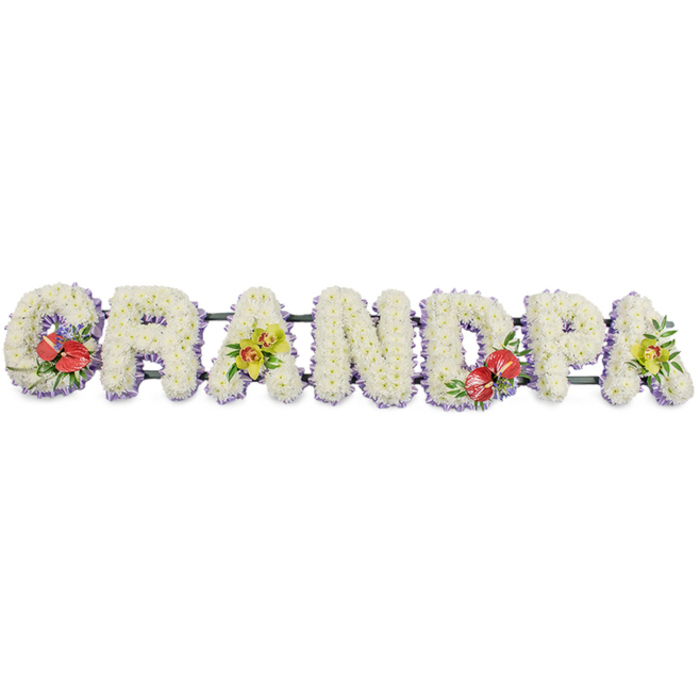 A dignified floral display stretches horizontally, spelling the word "GRANDPA" in strong block capitals, each letter standing proudly against a plain white backdrop that highlights every detail of the design. The letters are densely packed with small, pristine white chrysanthemums, their rounded petals forming a soft, textured blanket of blooms. Up close, the surface feels almost velvety, each flower head nestled snugly into the next so that no gaps of greenery show through. A delicate band of pale lavender ribbon sits neatly along the bottom of each letter, like a gentle lilac shadow that outlines the word and introduces a calm, soothing hue. All seven letters sit on a discreet dark green support rail, keeping the tribute straight and secure for display at a funeral service, wake, or chapel of rest in East Finchley or the surrounding North London area. Carefully placed accent sprays break the expanse of white with meaningful colour. On the "G" and "D", striking clusters of deep red anthuriums emerge, their glossy, heart-shaped petals and prominent central spadices shining against fresh green foliage and tiny touches of purple filler blooms. These bold, passionate reds contrast beautifully with the purity of the chrysanthemums. On the first and last "A", graceful sprays of yellow-green orchids, likely cymbidium, spill out elegantly from the letters, their arching stems and waxy petals symbolising enduring beauty and remembrance. The arrangement feels personal and heartfelt, a bespoke funeral tribute created to honour a beloved grandfather, perhaps someone who once walked East Finchley's quiet streets or enjoyed afternoons in nearby parks. Every flower and ribbon has been thoughtfully chosen, turning a simple word into a touching visual expression of love, respect, and gratitude.